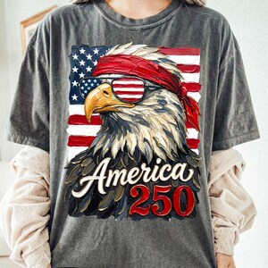 Puede incluir: Camiseta gris con una ilustraci&oacute;n detallada de un &aacute;guila calva americana con gafas de sol y una bandana roja, sobre un fondo de la bandera americana. El texto "America 250" se muestra debajo.
