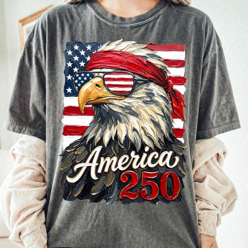 Puede incluir: Camiseta gris con una ilustraci&oacute;n detallada de un &aacute;guila calva americana con gafas de sol y una bandana roja, sobre un fondo de la bandera americana. El texto "America 250" se muestra debajo.