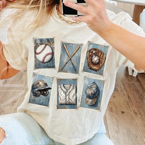 May include: Cream-colored t-shirt featuring a baseball-themed graphic. The design includes images of a baseball, crossed bats, a glove, a helmet, a jersey, and a cap, all set against a blue background.