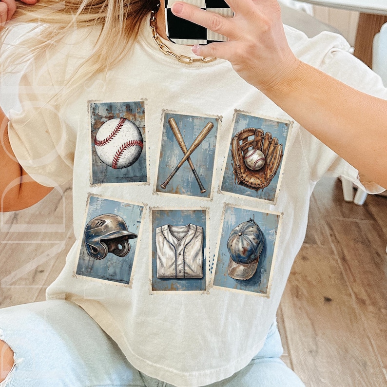May include: Cream-colored t-shirt featuring a baseball-themed graphic. The design includes images of a baseball, crossed bats, a glove, a helmet, a jersey, and a cap, all set against a blue background.