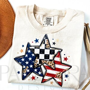 May include: Cream-colored t-shirt featuring three star designs with patriotic patterns. One star has a blue field with white stars, another has a black and white checkered pattern, and the third has red and white stripes. The shirt has a Comfort Colors tag.
