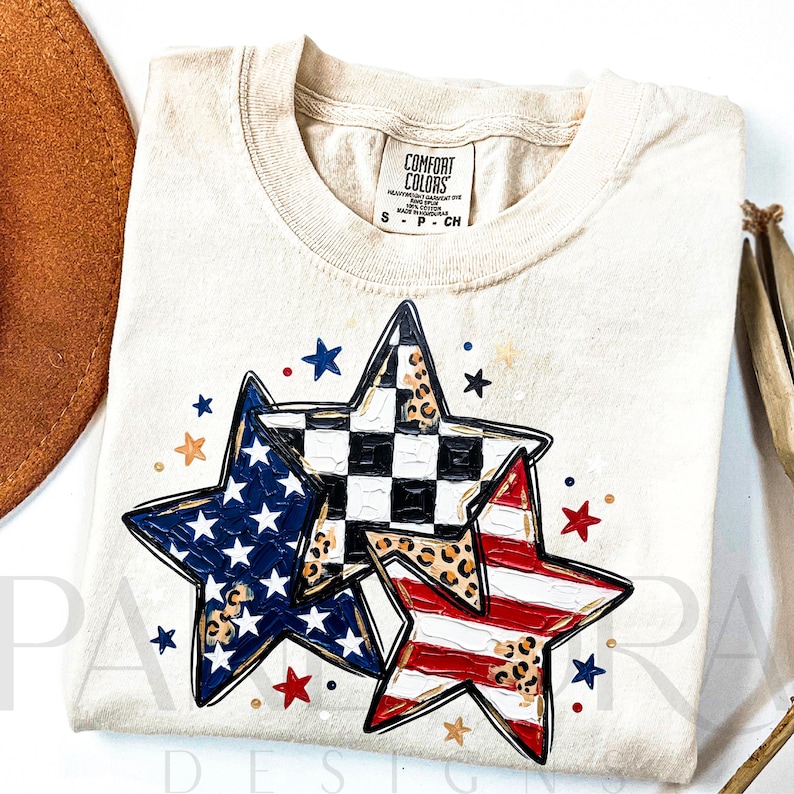 May include: Cream-colored t-shirt featuring three star designs with patriotic patterns. One star has a blue field with white stars, another has a black and white checkered pattern, and the third has red and white stripes. The shirt has a Comfort Colors tag.