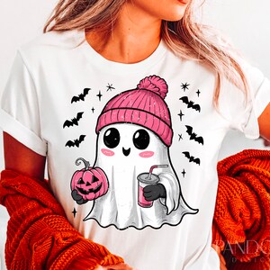 May include: White t-shirt featuring a cute cartoon ghost wearing a pink beanie hat, holding a pumpkin and a drink. The ghost has large eyes and a happy expression. There are bats and stars in the background.