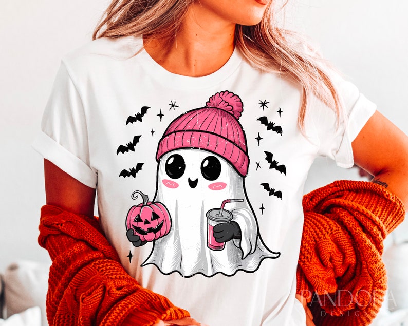May include: White t-shirt featuring a cute cartoon ghost wearing a pink beanie hat, holding a pumpkin and a drink. The ghost has large eyes and a happy expression. There are bats and stars in the background.
