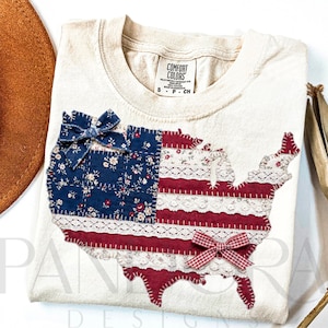 May include: Cream-colored t-shirt featuring a patchwork design of the United States in red, white, and blue fabrics. The design includes a blue floral fabric, red stripes, and white lace accents, with two small bows. The shirt has a Comfort Colors tag.