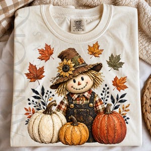 May include: A cream-colored t-shirt featuring a graphic of a scarecrow wearing a brown hat with a sunflower, a plaid shirt, and dark overalls. The design includes three pumpkins in white, orange, and rust, surrounded by autumn leaves and branches.