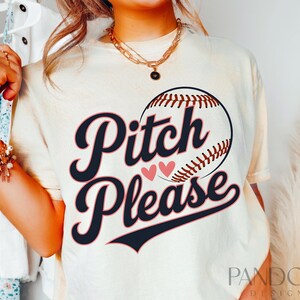 K&ouml;nnte beinhalten: Ein wei&szlig;es T-Shirt mit einem Baseball-Motiv und dem Text "Pitch Please" in Schwarz mit rosa Herzen. Das Shirt wird von einer Person mit einer goldenen Halskette getragen.