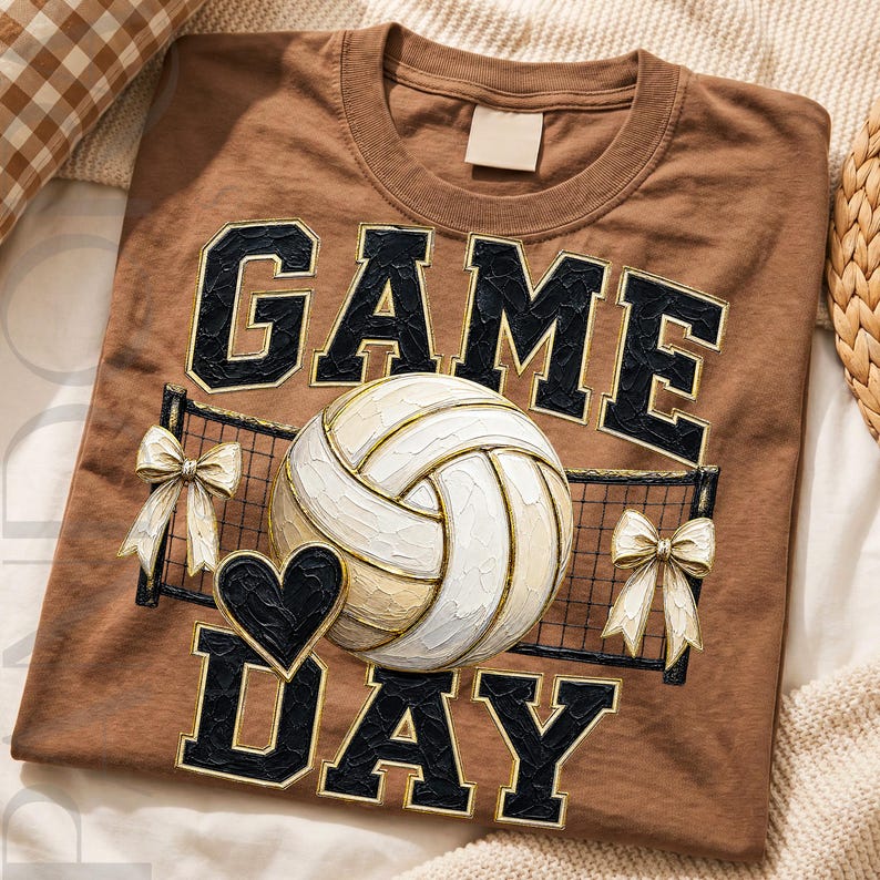 May include: A brown t-shirt featuring the text GAME DAY in black block letters with a white outline. The design includes a central white volleyball, a black heart, a black volleyball net graphic, and two cream-colored bows on either side.