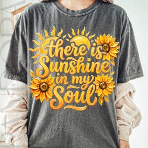 May include: Dark gray t-shirt with a vibrant yellow and orange graphic. The design features a sun, sunflowers, and the text "there is Sunshine in my Soul" in a flowing script. The shirt has short sleeves.
