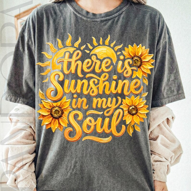May include: Dark gray t-shirt with a vibrant yellow and orange graphic. The design features a sun, sunflowers, and the text "there is Sunshine in my Soul" in a flowing script. The shirt has short sleeves.