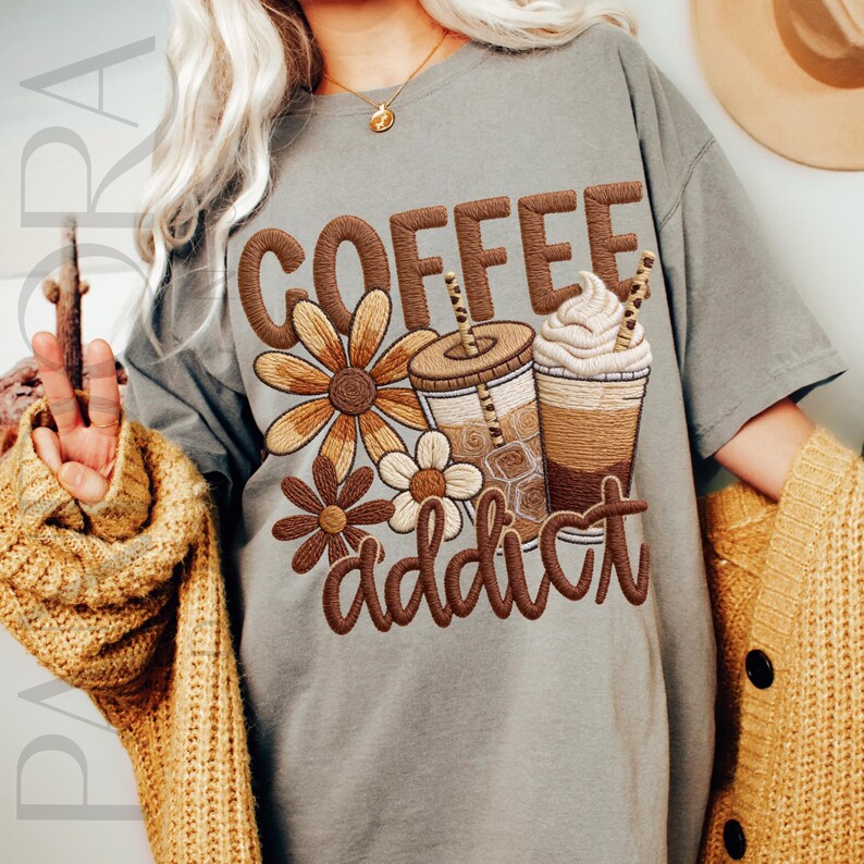 May include: A gray t-shirt with the embroidered words "COFFEE addict" and floral and coffee cup designs in brown. The shirt has short sleeves and a relaxed fit, perfect for casual wear.