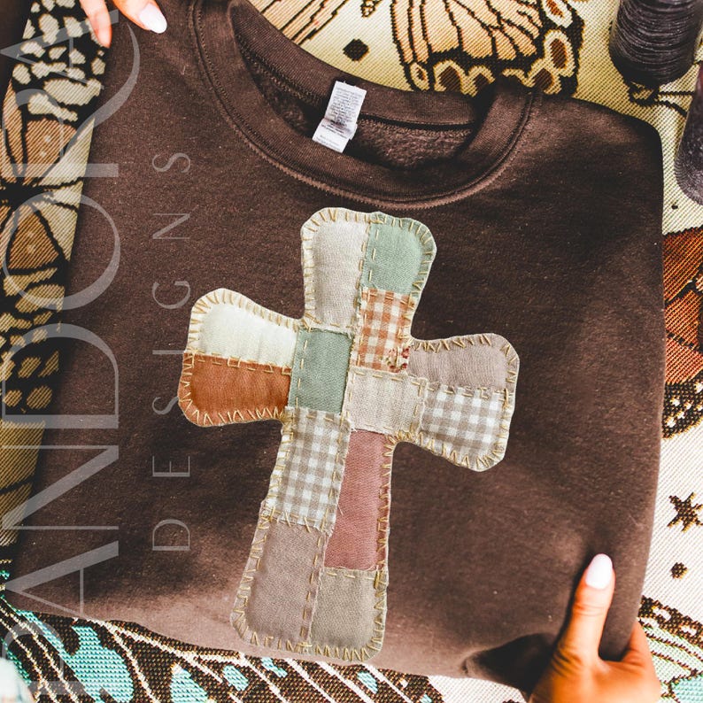 Peut inclure: Un sweat-shirt marron avec un motif de croix en patchwork. La croix est compos&eacute;e de carr&eacute;s de tissu vari&eacute;s dans les tons beige, vert et rose, cousus ensemble avec une couture d&eacute;corative. Le texte "AND DOT DESIGNS" est visible sur le c&ocirc;t&eacute; gauche du sweat-shirt.