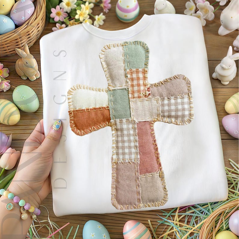 Peut inclure: Un t-shirt blanc &agrave; manches longues avec une croix patchwork aux couleurs pastel. La croix est compos&eacute;e de carr&eacute;s de tissu beige, vert et rose. Des d&eacute;corations de P&acirc;ques, dont des &oelig;ufs et des lapins, entourent le t-shirt.