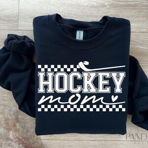 May include: Black sweatshirt with white text that reads "Hockey Mom" with a small heart and a hockey stick graphic. The text is surrounded by a checkered pattern.
