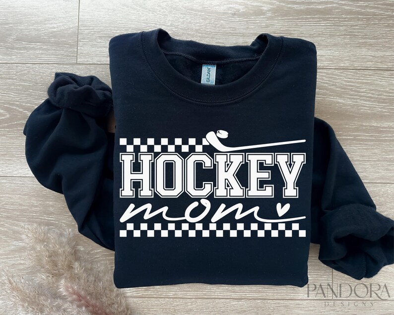 May include: Black sweatshirt with white text that reads "Hockey Mom" with a small heart and a hockey stick graphic. The text is surrounded by a checkered pattern.
