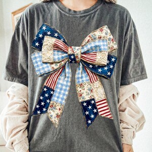 Pu&ograve; includere: T-shirt grigio con un grande motivo a fiocco. Il fiocco &egrave; un patchwork di motivi tra cui quadretti blu, strisce rosse, stampe floreali e stelle su sfondo blu navy. Il design del fiocco incorpora colori ed elementi patriottici.