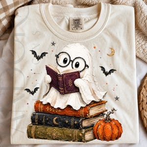 Peut inclure: Un t-shirt couleur crème avec un motif représentant un fantôme blanc portant des lunettes rondes noires et lisant un livre violet. Le fantôme est assis sur une pile de trois livres anciens orange, noirs et verts, à côté d'une citrouille orange.