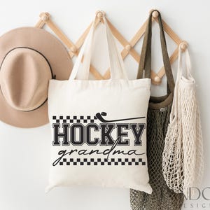 May include: White canvas tote bag with black text that reads "Hockey Grandma" with a black and white checkered pattern above and below the text. The bag is hanging on a wooden rack with a brown hat and two beige mesh bags.