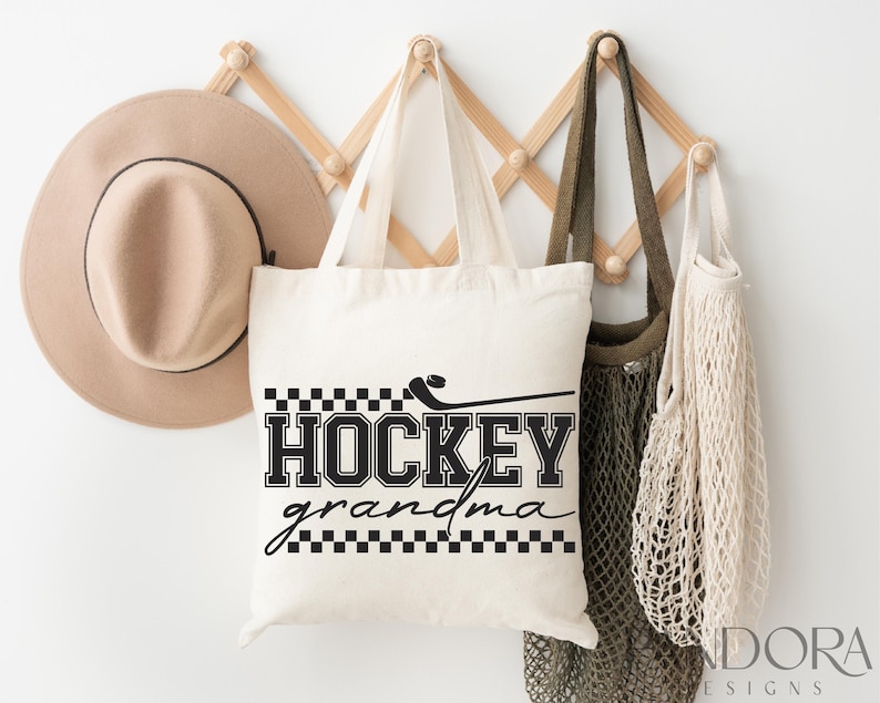 May include: White canvas tote bag with black text that reads "Hockey Grandma" with a black and white checkered pattern above and below the text. The bag is hanging on a wooden rack with a brown hat and two beige mesh bags.