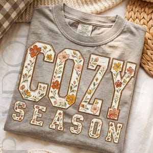 Peut inclure: Un t-shirt gris à col rond avec le texte COZY SEASON en grandes lettres majuscules. Les lettres sont blanches avec un contour marron et remplies d'un motif floral détaillé dans des tons orange, jaune et vert. Le t-shirt porte une étiquette Comfort Colors.