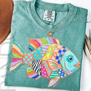 May include: A seafoam green t-shirt featuring a colorful, patchwork-style fish design. The fish has a blue face, and its body is made up of various patterns and colors, including pink, orange, yellow, and blue. A gold necklace with a small pendant is on the shirt.