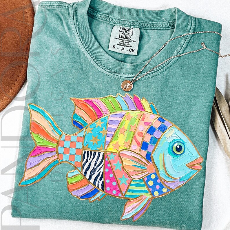 May include: A seafoam green t-shirt featuring a colorful, patchwork-style fish design. The fish has a blue face, and its body is made up of various patterns and colors, including pink, orange, yellow, and blue. A gold necklace with a small pendant is on the shirt.