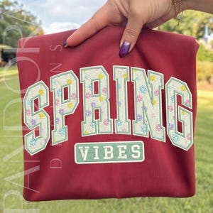 May include: A maroon sweatshirt with the word "SPRING" in large, embroidered letters filled with pastel floral patterns. Below, the word "VIBES" is embroidered in a rectangular box. The sweatshirt is held up by a hand.