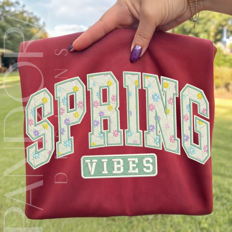 May include: A maroon sweatshirt with the word "SPRING" in large, embroidered letters filled with pastel floral patterns. Below, the word "VIBES" is embroidered in a rectangular box. The sweatshirt is held up by a hand.