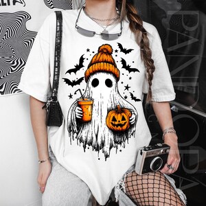 May include: White t-shirt with a Halloween ghost graphic. The ghost is wearing an orange beanie and holding a pumpkin and a drink. Black bats and stars are around the ghost. The design is in black and orange.