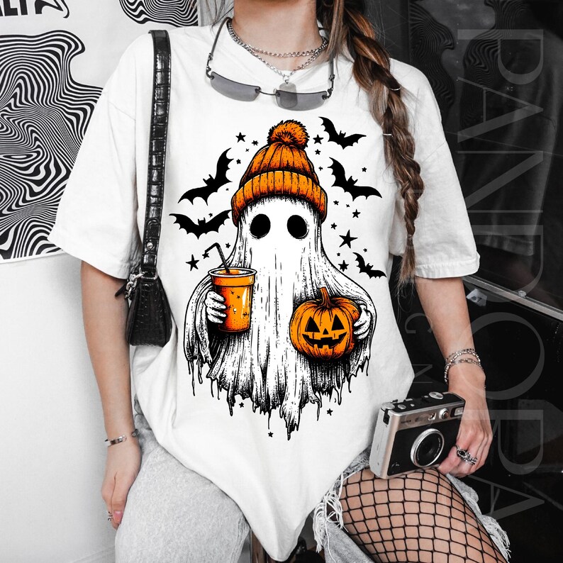 May include: White t-shirt with a Halloween ghost graphic. The ghost is wearing an orange beanie and holding a pumpkin and a drink. Black bats and stars are around the ghost. The design is in black and orange.
