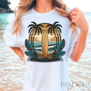 May include: White t-shirt with a graphic design of a surfboard in front of a sunset with palm trees and ocean waves.