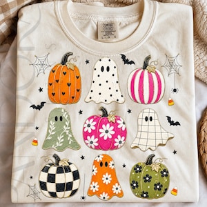May include: A cream-colored t-shirt featuring a graphic print of various Halloween-themed elements. The design includes pumpkins with heart, daisy, and checkered patterns, ghosts with polka dot, floral, and grid designs, plus bats, spiderwebs, and candy corn.