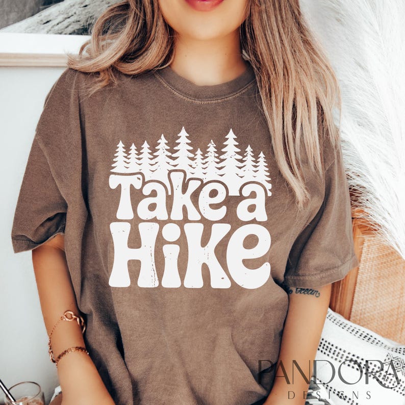 Może przedstawiać: Brązowa koszulka z białym nadrukiem graficznym z napisem "Take a Hike" z linią drzew za tekstem.
