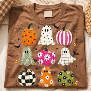 Op de afbeelding: Een bruin t-shirt met een grafische print van diverse pompoenen en geesten. De pompoenen tonen patronen zoals hartjes, bloemen en ruitjes. De geesten zijn versierd met stippen, bloemranken en rasters, omringd door vleermuizen en spinnenwebben.