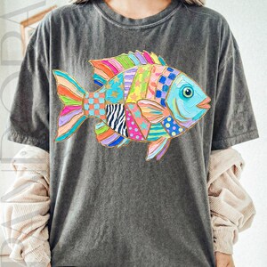 May include: Dark gray t-shirt featuring a colorful, patchwork-style fish design. The fish has a variety of patterns and colors, including blue, orange, pink, and green. The shirt is a casual, comfortable style.
