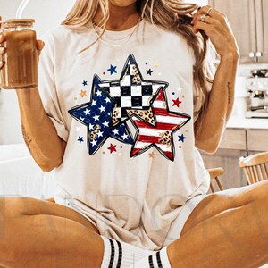 May include: A cream-colored t-shirt featuring three star designs. One star has a blue and white pattern, another has a red and white striped pattern, and the third has a black and white checkered pattern with a leopard print. The shirt has a round neckline and short sleeves.