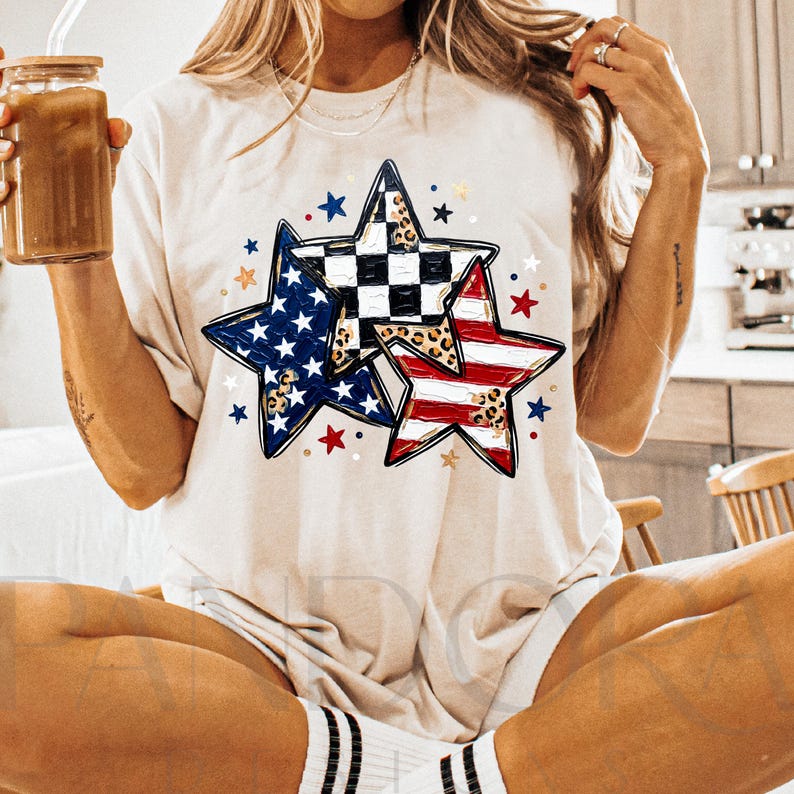 May include: A cream-colored t-shirt featuring three star designs. One star has a blue and white pattern, another has a red and white striped pattern, and the third has a black and white checkered pattern with a leopard print. The shirt has a round neckline and short sleeves.