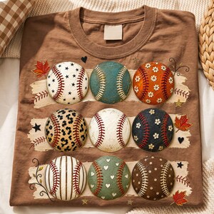 Op de afbeelding: Een bruin t-shirt met een grafische print van negen honkballen in drie rijen. Elke honkbal heeft een uniek patroon, waaronder sterren, bloemen, luipaardprint, strepen, hartjes en stippen, geaccentueerd door herfstbladeren.