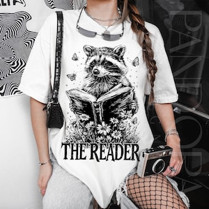 Puede incluir: Camiseta blanca con una ilustración en blanco y negro de un mapache leyendo un libro, rodeado de mariposas y flores. El texto "THE READER" está impreso debajo. La camiseta tiene mangas cortas y un corte holgado.