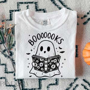 May include: White t-shirt with a black and white illustration of a ghost reading a book. The text "BOO0000KS" is above the ghost.