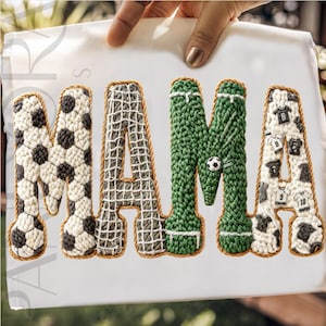 May include: A white sheet with the word "MAMA" in large, textured letters. The letters are decorated with soccer ball patterns, netting, and green crochet, with a small soccer ball and jersey graphics.