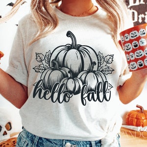 Peut inclure: Un t-shirt blanc avec un motif noir de citrouilles et de feuilles. Le texte "hello fall" est &eacute;crit sous les citrouilles.