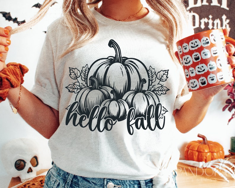Peut inclure: Un t-shirt blanc avec un motif noir de citrouilles et de feuilles. Le texte "hello fall" est &eacute;crit sous les citrouilles.