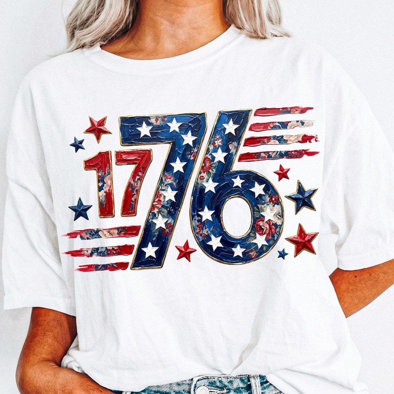 May include: White t-shirt featuring a patriotic design with the number "1776" in red, white, and blue. The "76" is filled with a floral pattern and surrounded by stars. Red and blue brushstroke accents complete the design.