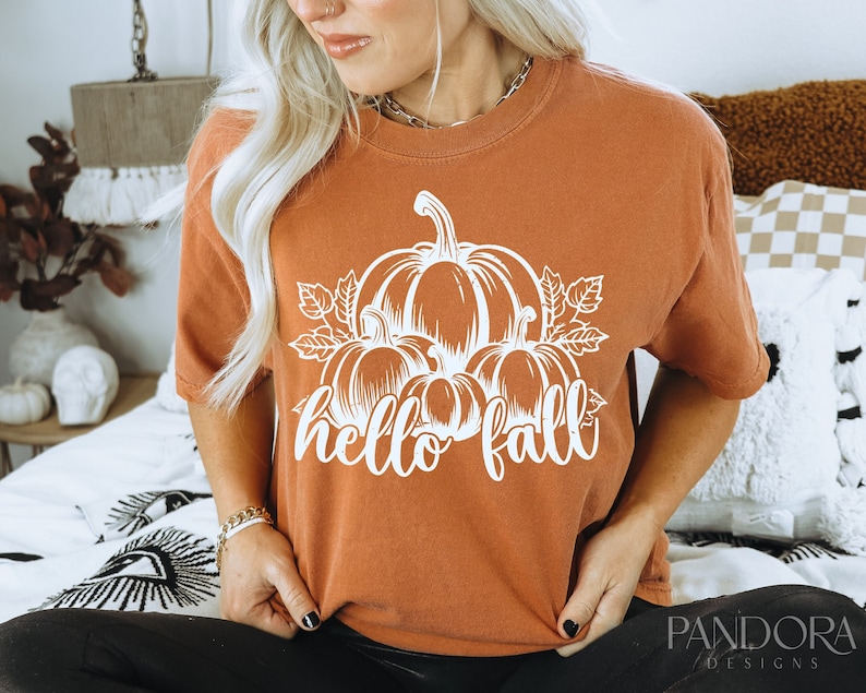Peut inclure: Un t-shirt couleur rouille avec un motif blanc de citrouilles et le texte "hello fall".