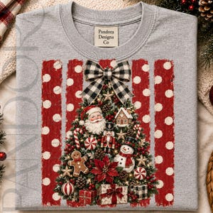 Puede incluir: Una camiseta gris jaspeado de cuello redondo con un gráfico de un árbol de Navidad decorado con una cara de Santa Claus, un cascanueces, un hombre de jengibre, un muñeco de nieve y regalos. Un lazo de cuadros blanco y negro corona el árbol, centrado sobre rayas rojas con lunares blancos.