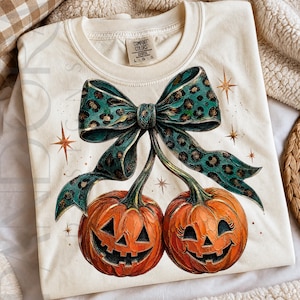 May include: Cream-colored sweatshirt featuring a Halloween-themed design. The graphic includes two orange jack-o'-lanterns with smiling faces, connected by a teal bow with a leopard print pattern. Gold starbursts accent the design.