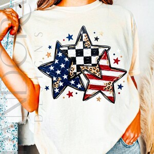 May include: Cream-colored t-shirt featuring three star designs with patriotic patterns. One star has a blue and white design, another has red and white stripes, and the third has a black and white checkered pattern with leopard print accents.