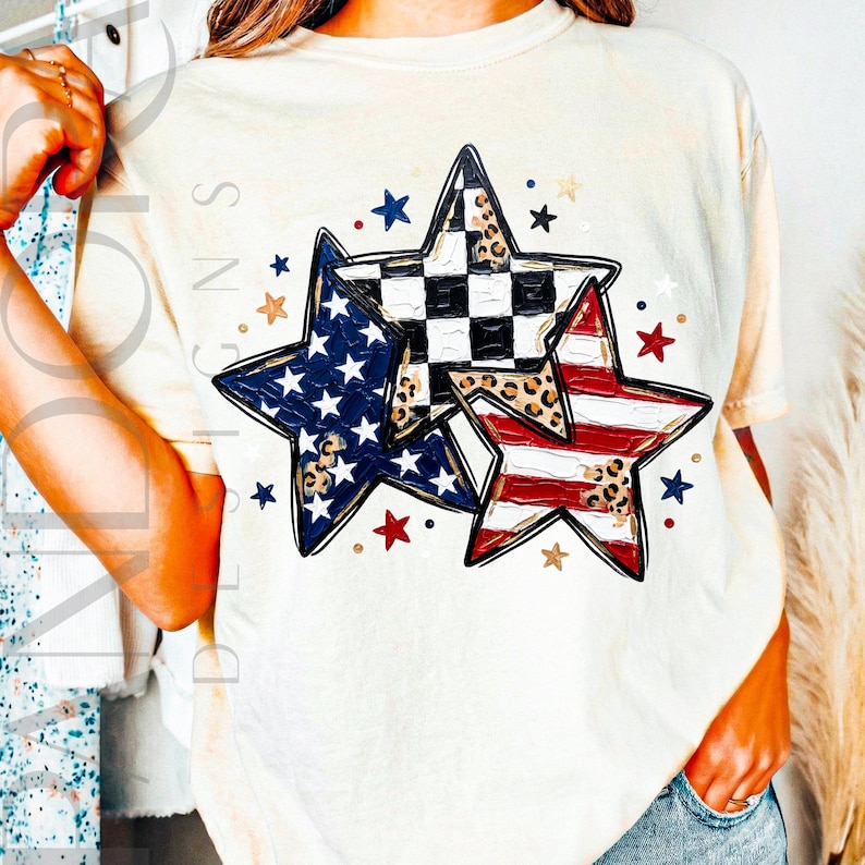 May include: Cream-colored t-shirt featuring three star designs with patriotic patterns. One star has a blue and white design, another has red and white stripes, and the third has a black and white checkered pattern with leopard print accents.
