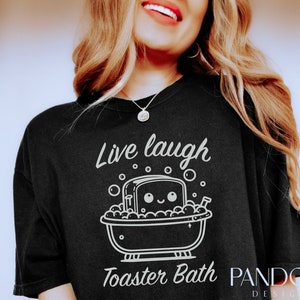K&ouml;nnte beinhalten: Ein schwarzes T-Shirt mit einer wei&szlig;en Grafik einer Scheibe Brot in einer Badewanne mit Blasen und dem Text "Live laugh Toaster Bath".